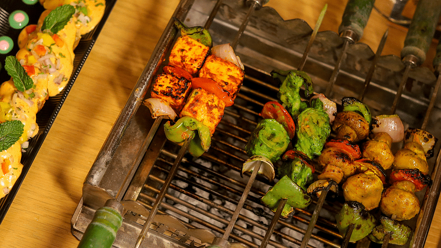 Various marinated kebabs cooked on skewers over a grill, accompanied by green bamboo handles - Themis Barbecue House, Ambience Mall, Gurugram