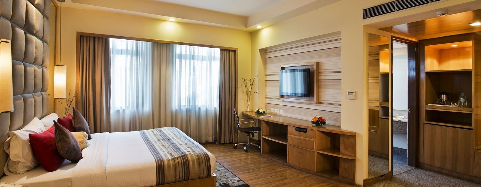 A sideview of a bed in a room with TV and a huge glass window | The Manor Bareilly