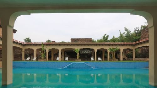 The swimming pool at Tijara Fort-Palace, a Neemrana property in Alwar, Rajasthan, is framed by arched walkways and lounge chairs under an overcast sky.