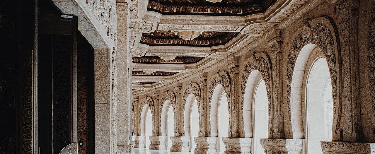 A luxurious hallway with arched windows, columns, and a polished marble floor, evoking a sense of grandeur and opulence.