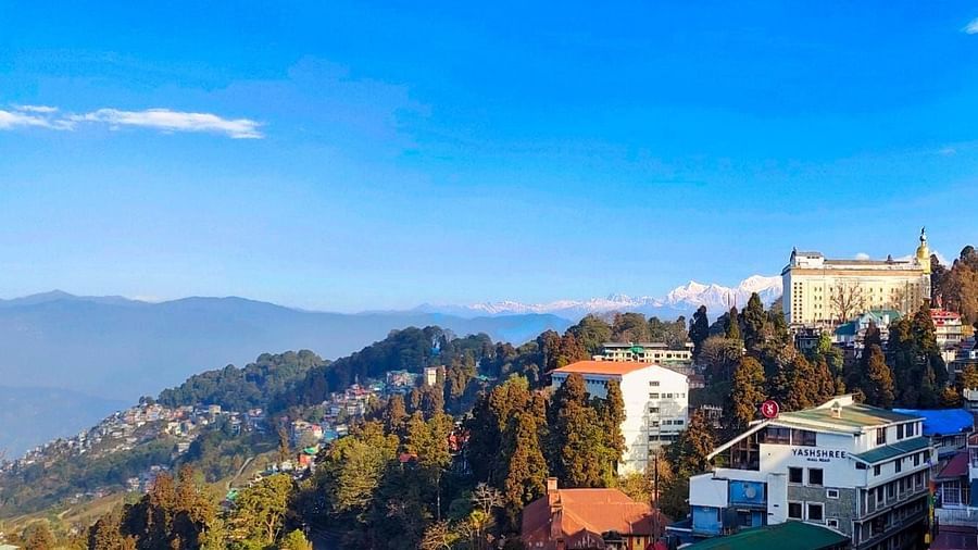 Stunning view of a hillside town with lush greenery, colourful rooftops, and snow-capped mountains in the background under a clear blue sky.