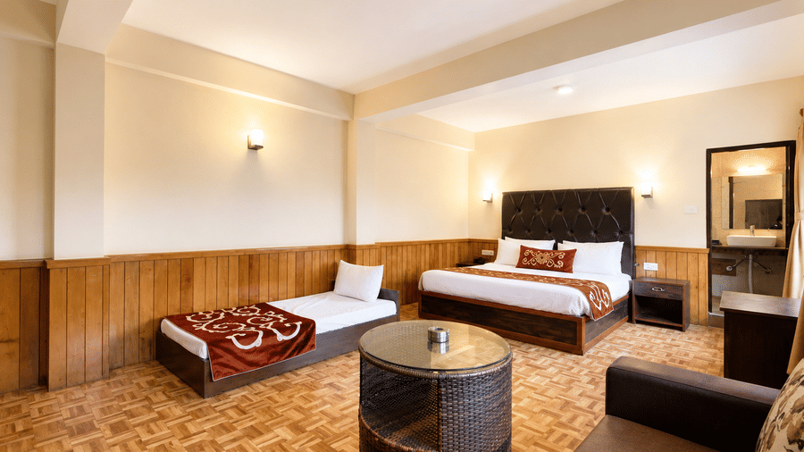 Premium Room at Orsino Khangri Karpo Retreat & Spa, Lachung contains a large bed with a dark tufted headboard. A single bed sits nearby, along with comfortable seating and wooden panelling.