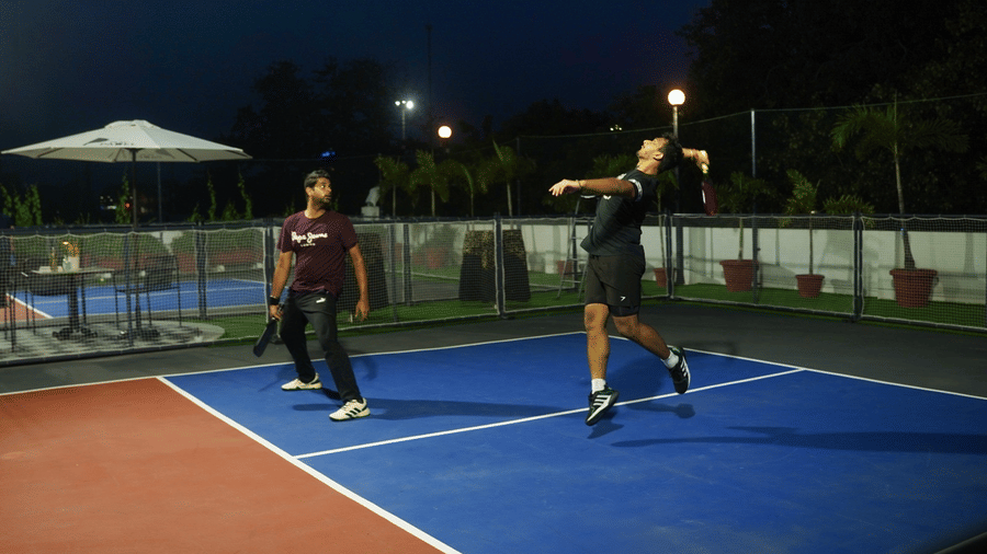 A night-time outdoor sports court with 2 people playing on a fenced hard court under floodlights at Clarks Avadh, Lucknow.