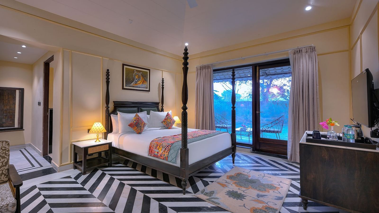 Luxury Suite Cottage featuring a four-poster bed with white linens, a colourful patterned runner and pillows, a patterned armchair, a wall-mounted TV, and sliding glass doors leading to a balcony - Singhvilas, Ranthambore