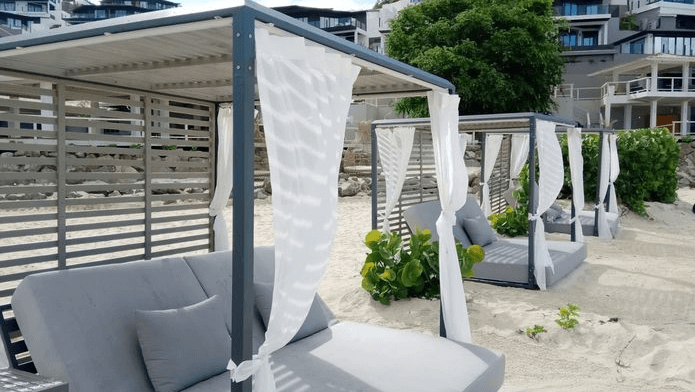 Luxurious beachfront daybeds with white canopies on a sandy area, resort buildings in background.