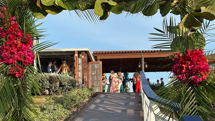An wooden pathway leading to the cottage style courtyard where people have gathered to celebrate a wedding. The entrance of the decorated with branches, leaves, and flowers. 