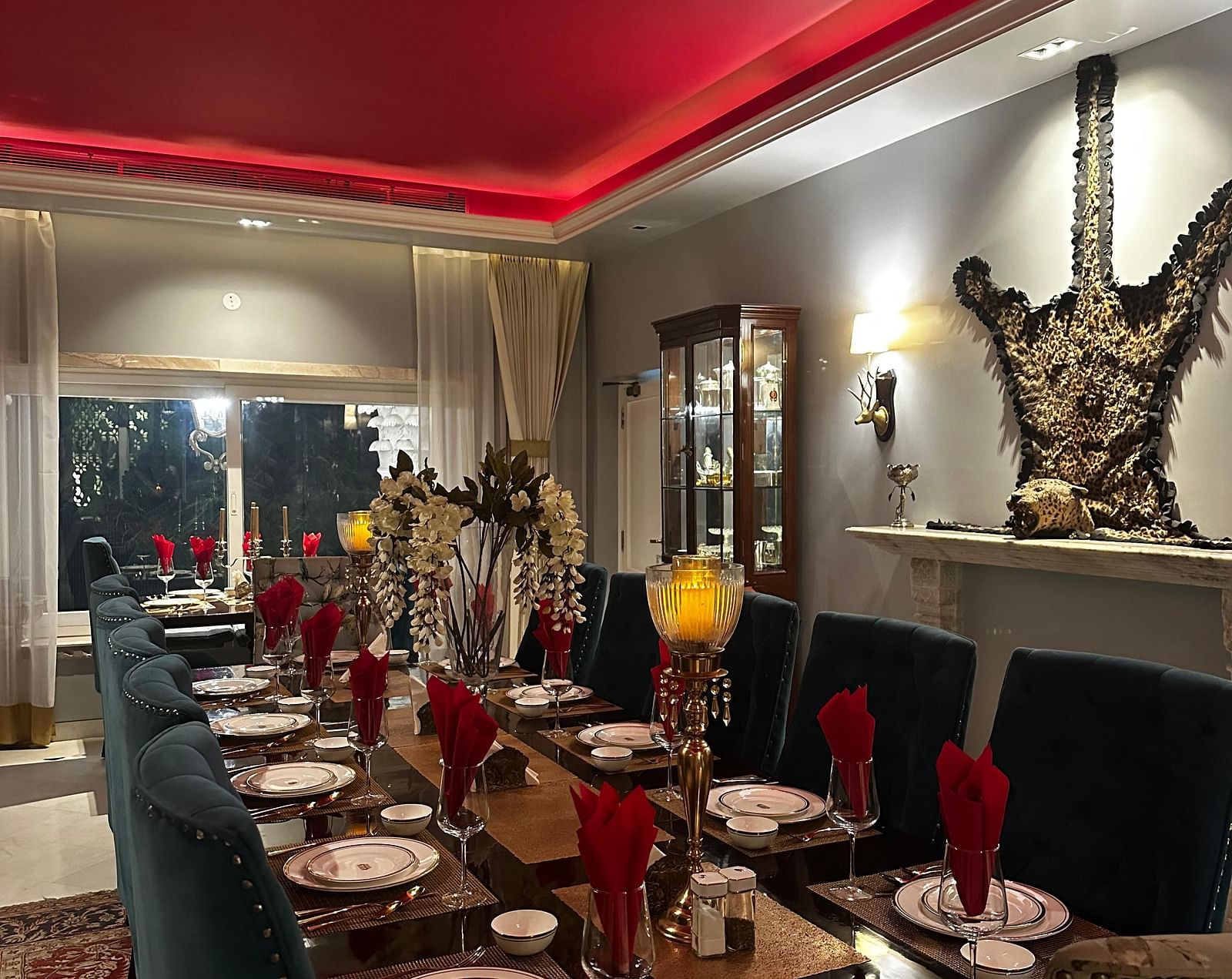 A long dining table set with plates and cutlery, under a red ceiling with neon lighting, in a room with grey walls and decorative wall sconces | Dileep  Kothi, Jaipur