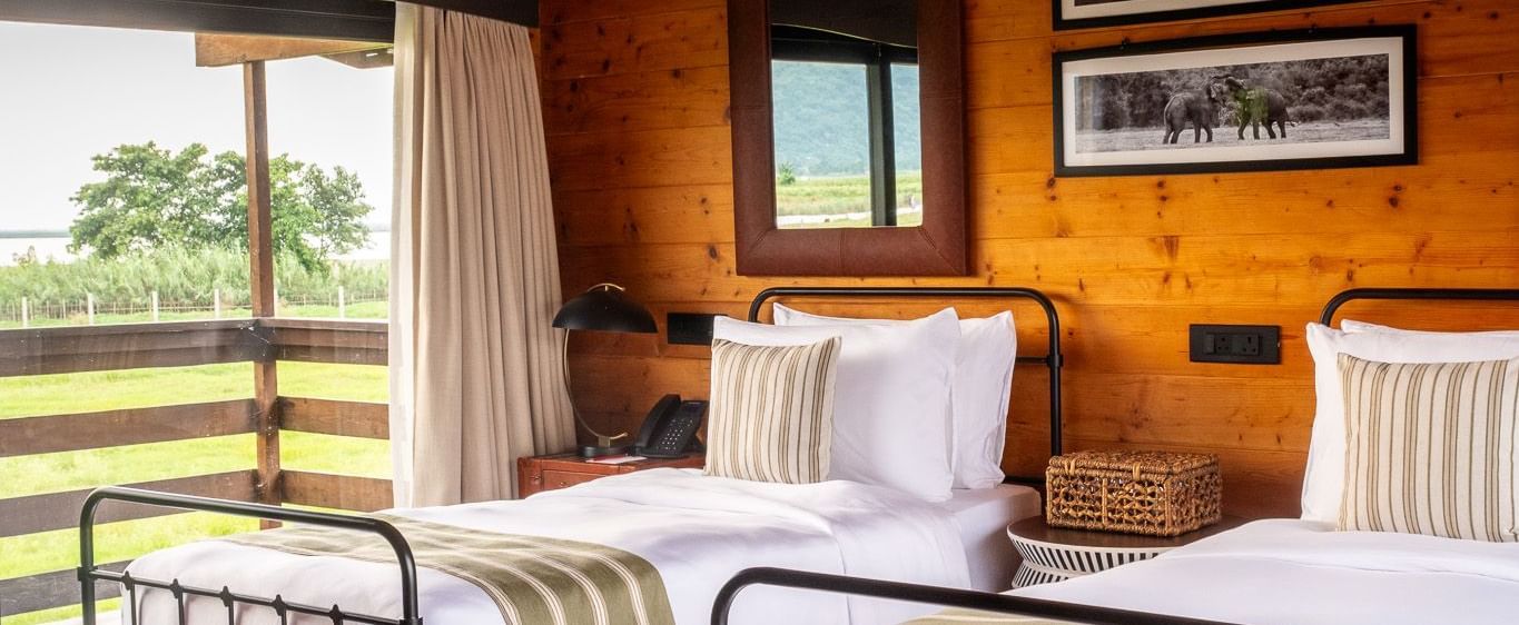 A bright and airy twin bedroom with two metal-framed beds, each with a white comforter and green striped runner. The room has wooden walls and a sloped ceiling. A large window provides a beautiful view of a grassy landscape and a body of water. This is a r