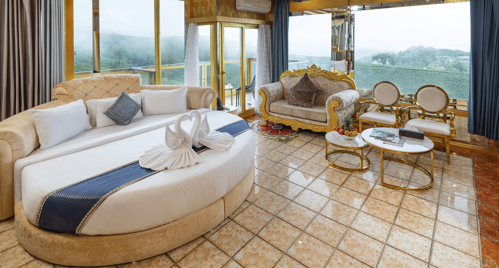 A wide-view of the Jacuzzi 180-degree Suite bedroom at Parakkat Nature Hotel and Resorts, Munnar, featuring decorative chairs and a sofa, patterned floor tiles, and large windows providing a view of outdoors.