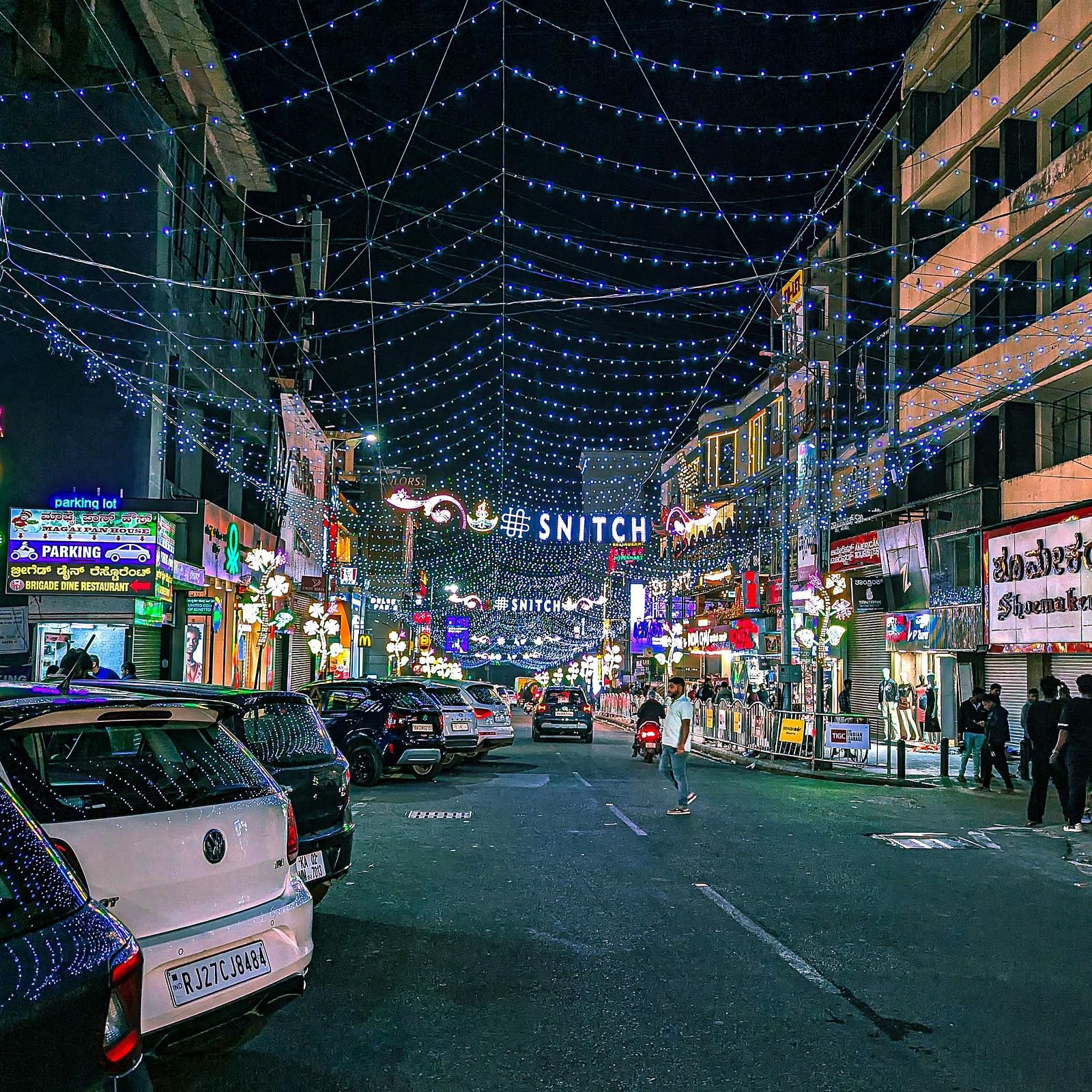 Nighttime scene of Brigade Road, Bangalore, illuminated with lights and featuring shops and vehicles.