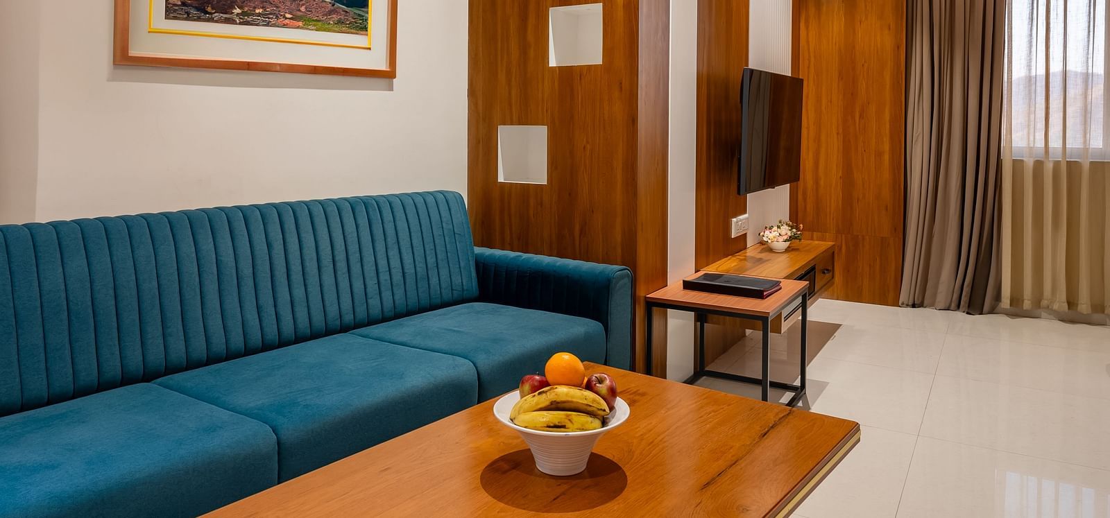 A hotel living area features a comfortable blue sofa, a wooden coffee table with fruit, and framed landscape artwork - Lords Eco Inn Ambaji.