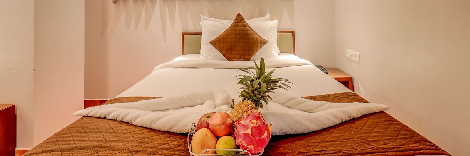 A spacious bed at Harigovinda's JNStay, with neatly arranged blankets, pillows, and a fruit basket, with windows covered by curtains above