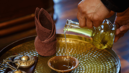A hand pouring oil into a small bowl, set on a golden table with other spa items.