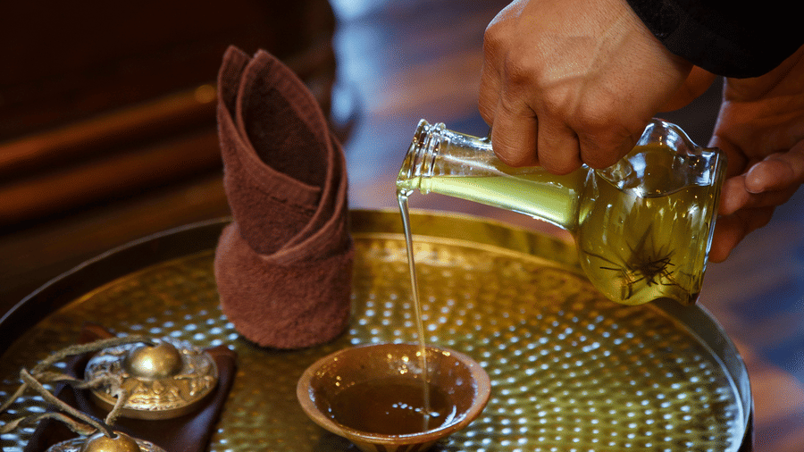 A hand pouring oil into a small bowl, set on a golden table with other spa items.