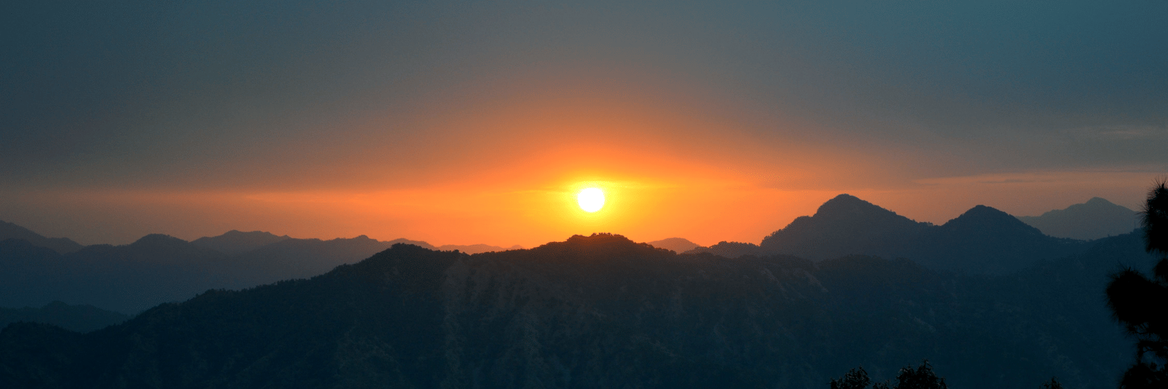 Sunset over the mountains of Kasauli, with the sun glowing above layered mountain peaks and silhouetted trees in the foreground.
