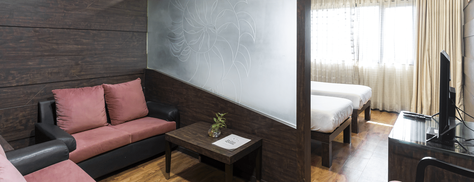 Room with sofa set, table, twin beds and large window with curtains at Infiniti Hotel, Indore.