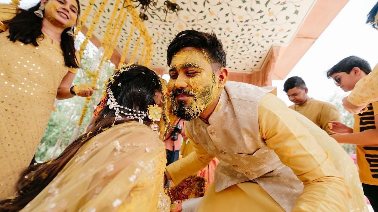 A groom with turmeric-covered face leans in playfully towards his bride during a joyful haldi ceremony, surrounded by loved ones.- Umaid Palace - A Lakeside Heritage Palace Resort