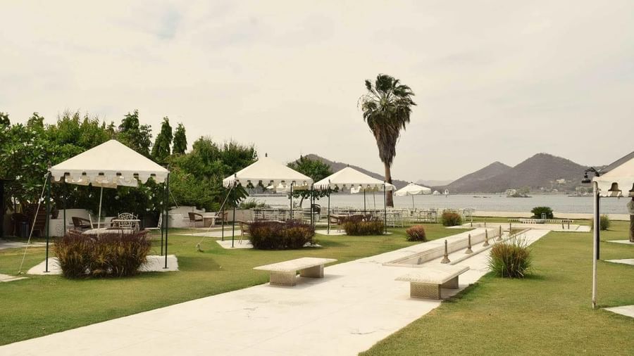 Outdoor seating area with white tents, benches, and lush green lawn by a lake with hills in the background at Ram Pratap Palace, Udaipur.
