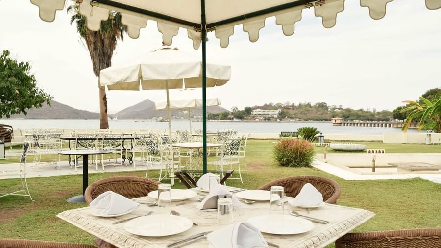 Outdoor dining setup at Ram Pratap Palace, Udaipur with a marble table, chairs, plates, and napkins under a canopy, overlooking the lake and distant hills.