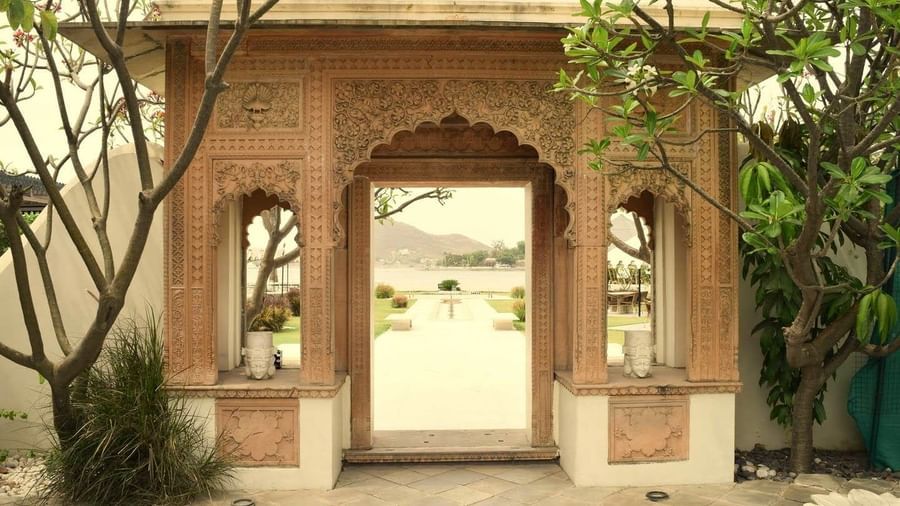 Ornate stone archway at Ram Pratap Palace, Udaipur framed with floral carvings and greenery, opening to a garden path with a lake and hills visible in the distance.