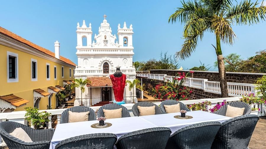 An outdoor dining area with a long table set for many guests, surrounded by white chairs, situated in front of a white church at Fort Tiracol, Goa with multiple bell towers.