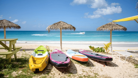 A couple of colorful kayaks and paddleboards on a sandy beach with thatched umbrellas near Tamarind Hills Resort and Villas, Antigua.