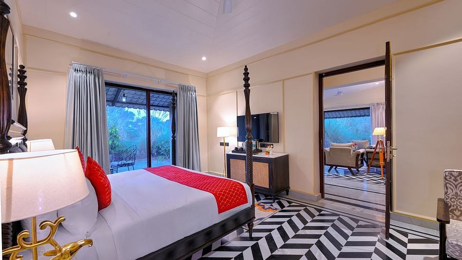 Premium Suite Cottage with a dark wood four-poster bed featuring red accents, black-and-white chevron flooring, a seating area, a wall-mounted TV, and an open doorway leading to another room - Singhvilas, Ranthambore