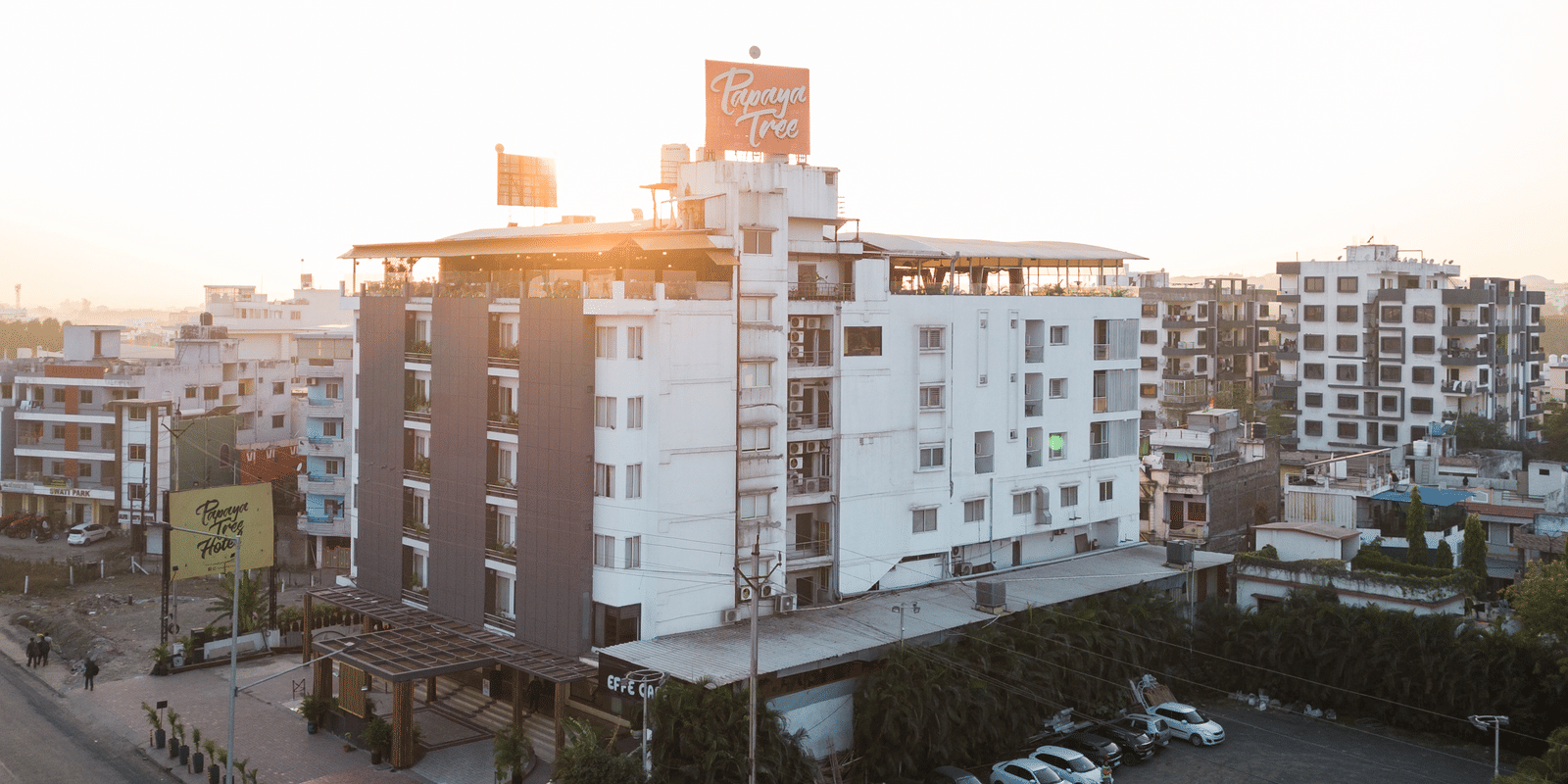 A wide exterior view of Papaya Tree Hotels captured during sunset, highlighting the building’s white facade, rooftop area, and surrounding city streets with parked vehicles.