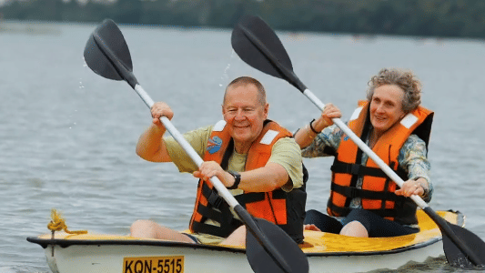 Two people kayaking on still backwaters, smiling as they paddle together while wearing safety gear and life jackets.