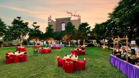 An outdoor event space with tables draped in red is set on a green lawn at dusk - Lion Lords Inn Rajula