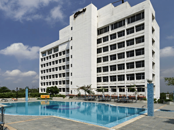 A large, modern white hotel building is shown behind a spacious blue swimming pool with lounge chairs on the patio - Clarks Avadh, Lucknow