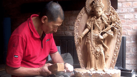 A stone carver working carefully beside a completed traditional Hindu deity statue in a historic masonry workshop in Patan, Nepal.