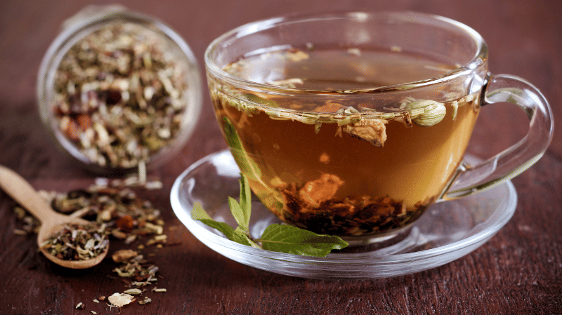 A glass cup filled with herbal tea, with loose-leaf tea floating inside and more loose-leaf tea spilled from a jar and wooden spoon on a wooden table.