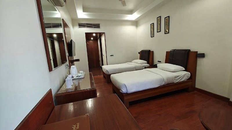 Corner view shot of a bedroom featuring two single beds along with dark wooden flooring with the entrance corridor at the far end of the room - Hotel Gold, Panipat