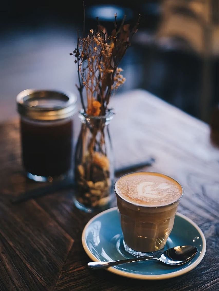A cup of cappuccino served on a blue coaster with a silver spoon on a wooden table with a flower vase beside it. 