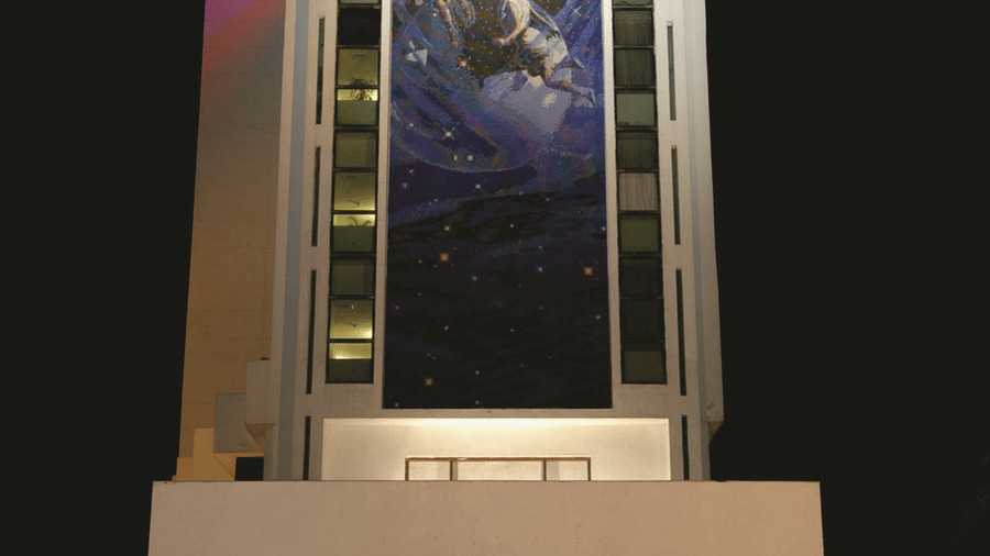 Hotel building exterior at night with a tall illuminated facade, a large banner display and a well-lit entrance with glass doors underneath.