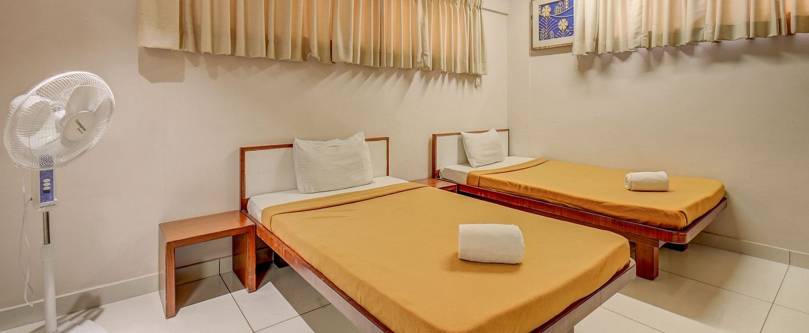 A spacious room at Harigovinda's JNStay, featuring two beds, a bedside table, two adjustable window panels covered with curtains, and a ceiling fan on the side