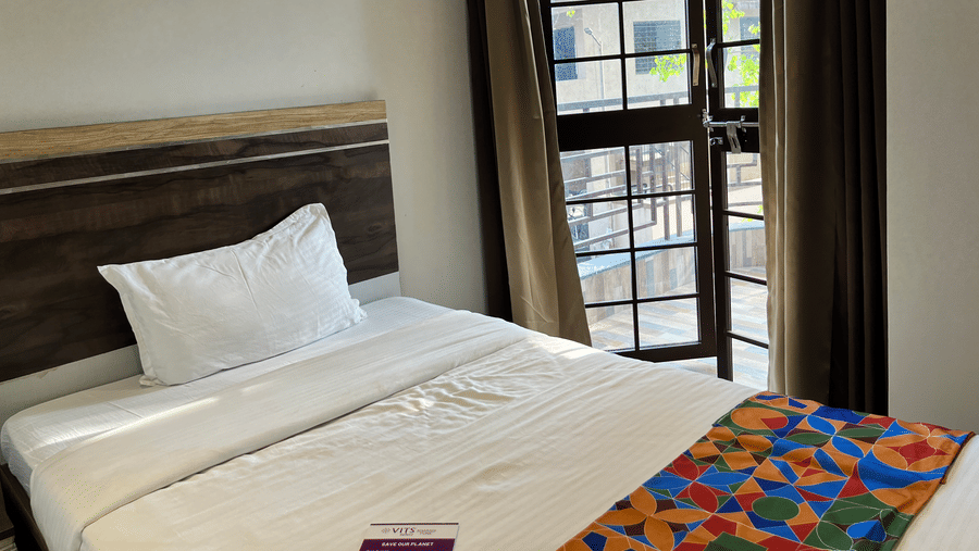 A close-up of a single bed near a glass & wooden door to the balcony, with green curtains next to the door inside Classic Single Room - the best hotel room in Kharadi at VITS Select Kharadi, Pune.