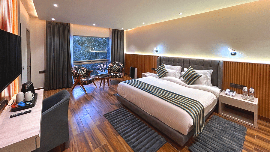 The premium room of DLS The Emerald, Nainital with a bed, bed side table, chairs, coffee table, a window overlooking the view, with warm lighting