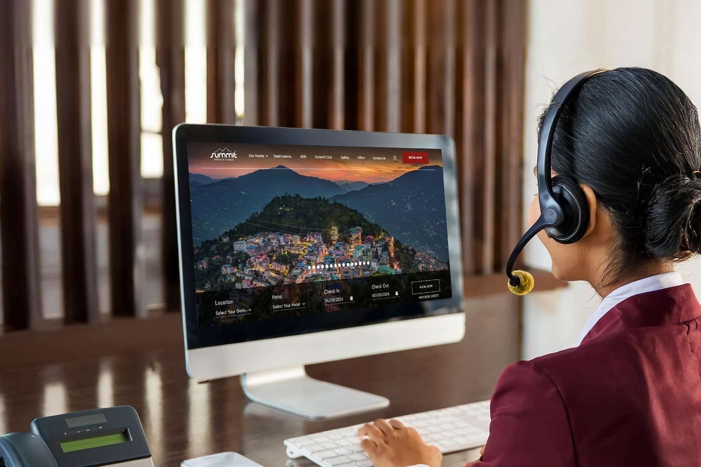 Female customer service representative in uniform using a headset and computer at a desk, assisting with hotel bookings on a website displaying a scenic town.