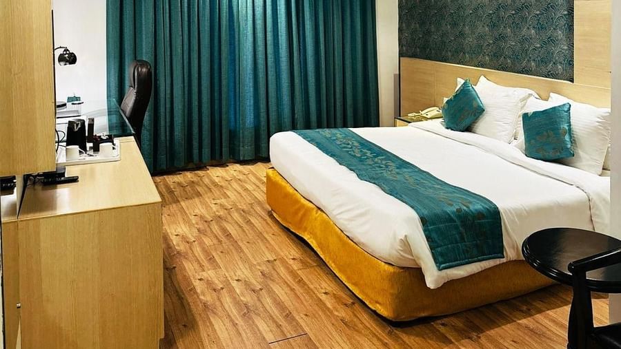 A cosy hotel room at Amer Greens featuring a large bed, hardwood floors, an accent wall, and a large window covered by floor-to-ceiling drapes. Room