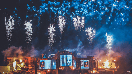 A far-out view of a stage inside Tomorrowland Thailand 2026 with many firecrackers bursting in the sky and people dancing to the songs in the concert.