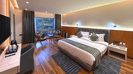 The premium room of DLS The Emerald, Nainital with a bed, bed side table, chairs, coffee table, a window overlooking the view, with warm lighting