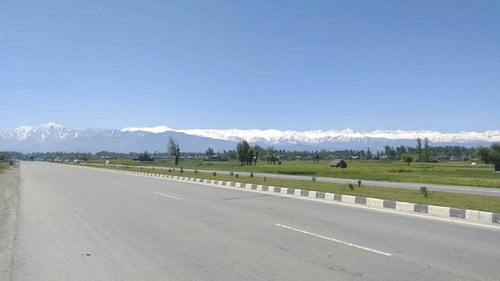 A wide road with lane markings, guardrails, open land on both sides, and distant mountains under a clear blue sky.