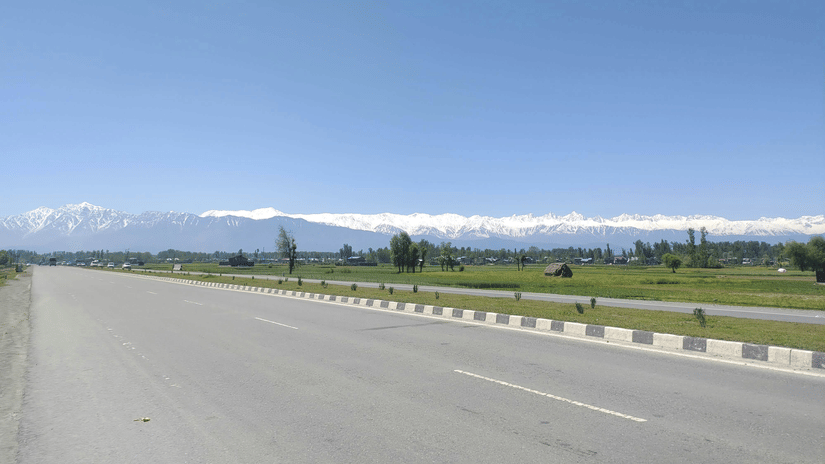 A wide road with lane markings, guardrails, open land on both sides, and distant mountains under a clear blue sky.