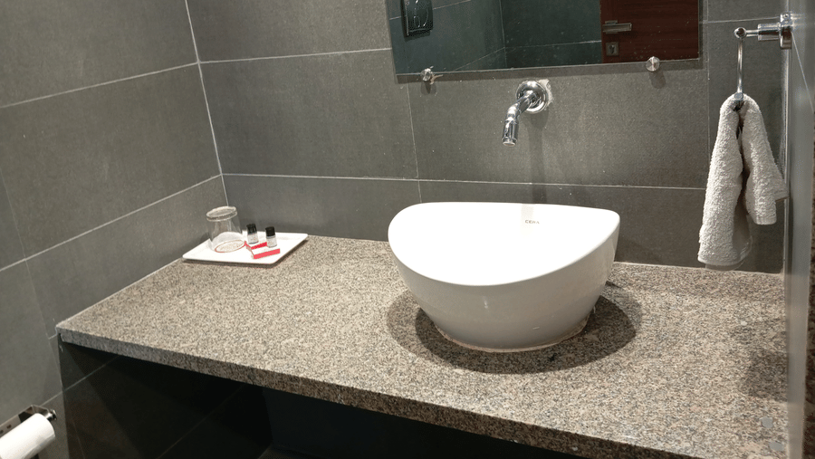 A modern bathroom at Hotel Ashray Inn Express featuring a countertop basin, wall mirror, towel holder, and tiled walls.