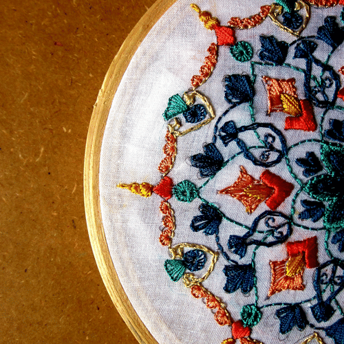 A close-up shot of a half-finished cross-stitch embroidery piece in a wooden hoop, featuring a colourful geometric pattern.