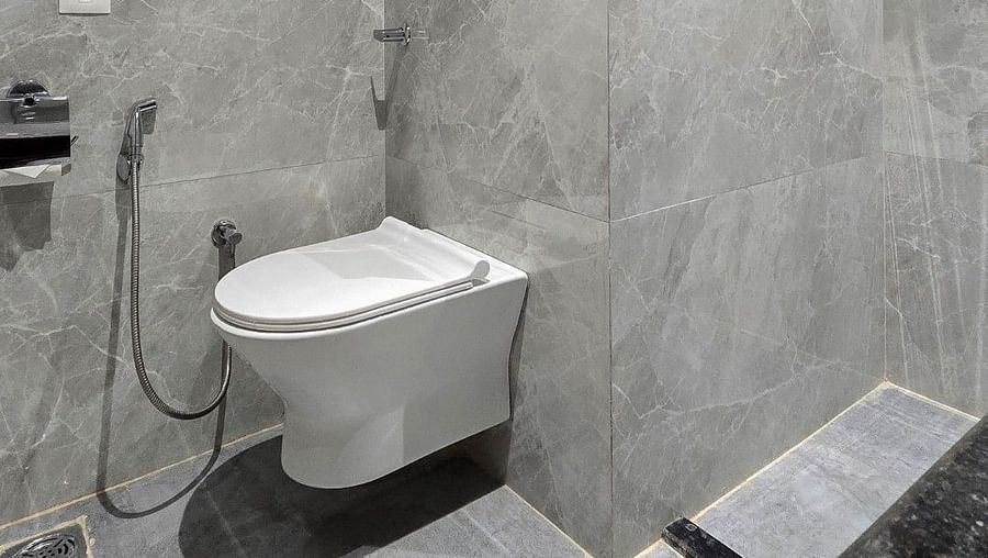 A hotel bathroom with a wall-mounted toilet against grey marble walls. Above the toilet, a towel rack holds neatly folded white towels. The lighting is soft and evenly distributed. - Clarks Inn Suites, Near Taj Mahal