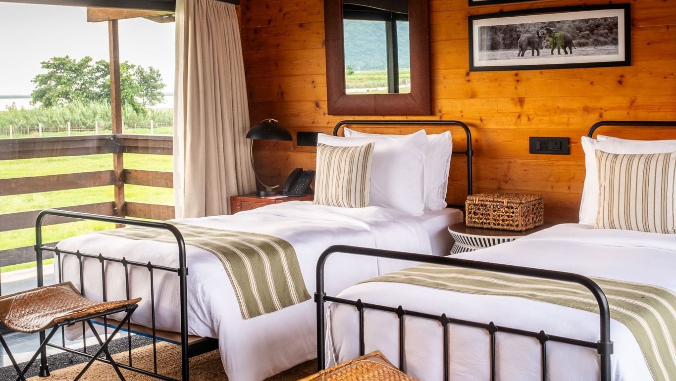 A bright and airy twin bedroom with two metal-framed beds, each with a white comforter and green striped runner. The room has wooden walls and a sloped ceiling. A large window provides a beautiful view of a grassy landscape and a body of water. This is a r