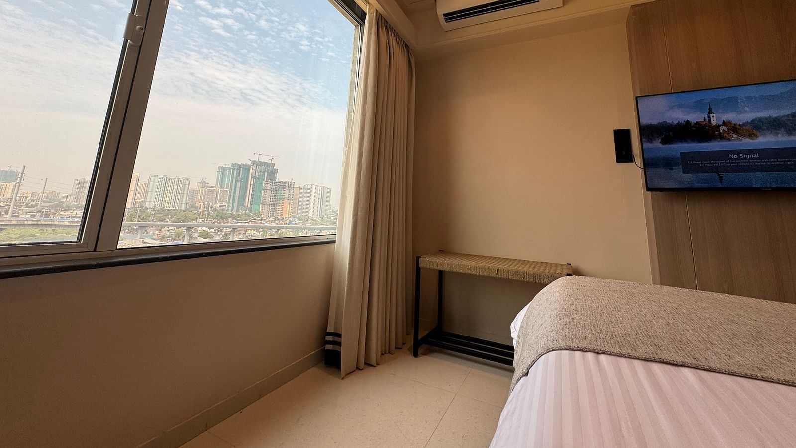 A city-view room with a bed and TV, simple decor, and a large window with a blue sky outside.